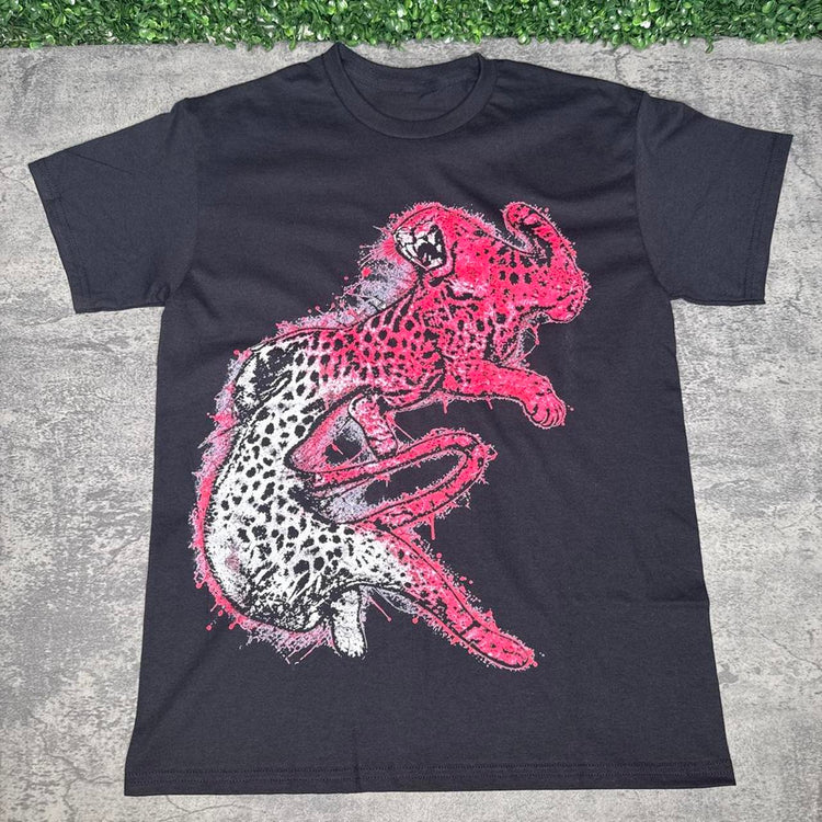 Black tee with a large pink-and-white spotted cheetah graphic on concrete with grass above