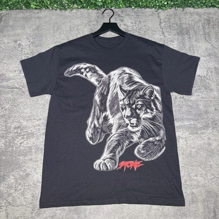 Black STONE panther tee on a hanger against gray concrete with green grass above