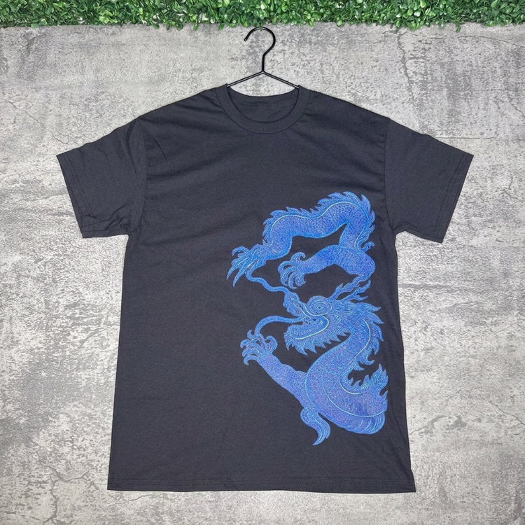 Black blue dragon tee hanging against gray concrete with green grass along the top