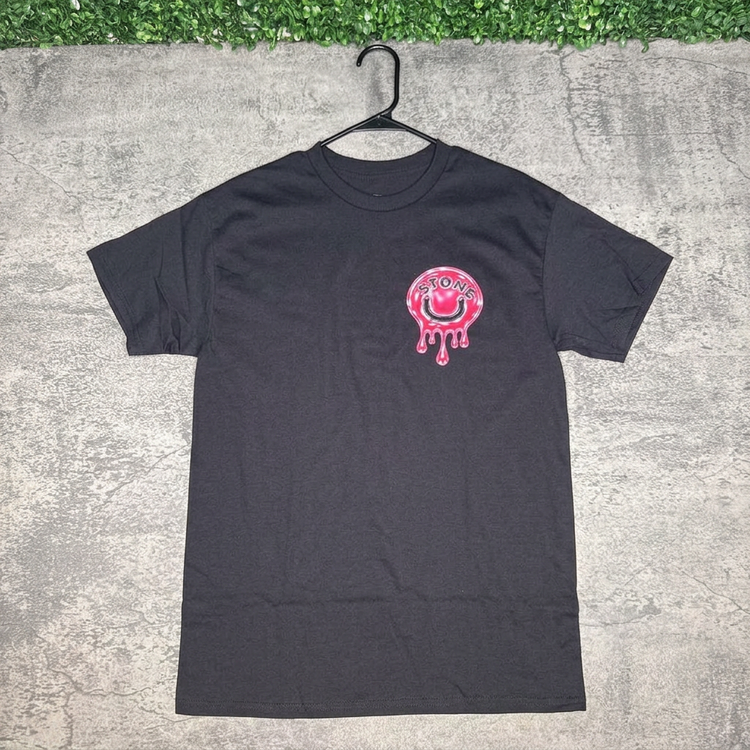 Black STONE pink drip smile tee hanging against concrete with green grass above